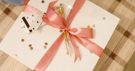 A glossy pink ribbon wraps horizontally and vertically around a white gift box. The ribbon forms a bow in the center.