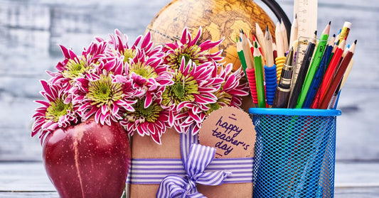 A teacher appreciation gift set with pink flowers, an apple, and a box wrapped in purple ribbon with a “Happy Teacher’s Day” tag, next to a globe and pencil holder.