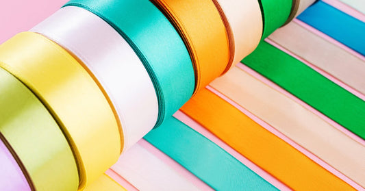 Rolls of satin ribbon in green, orange, white, turquoise, yellow, pink, and beige run diagonally on a pink background.