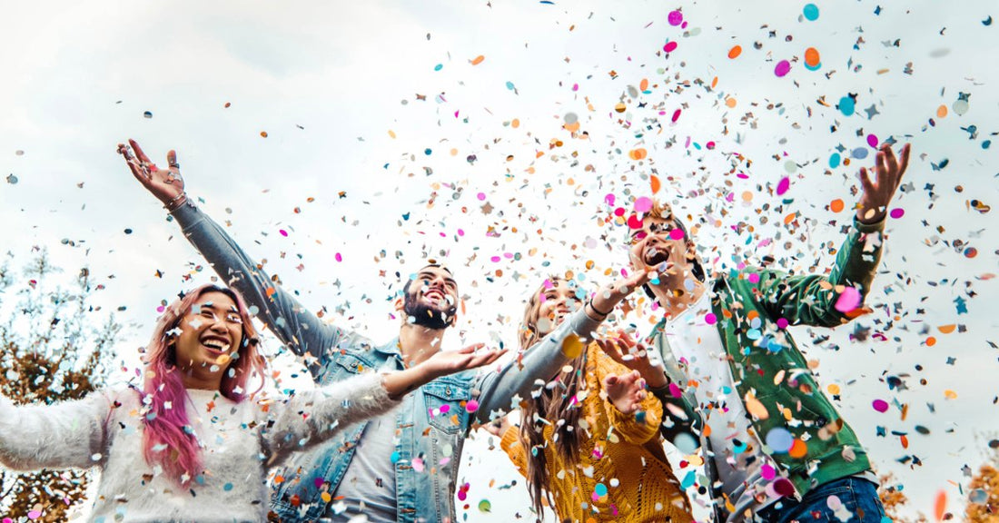 Upward view of four young adults standing outdoors with their arms raised beneath a shower of colorful confetti.