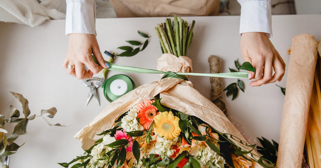 A florist ties a green ribbon around a bouquet of orange, yellow, pink, and white flowers wrapped in light brown paper.