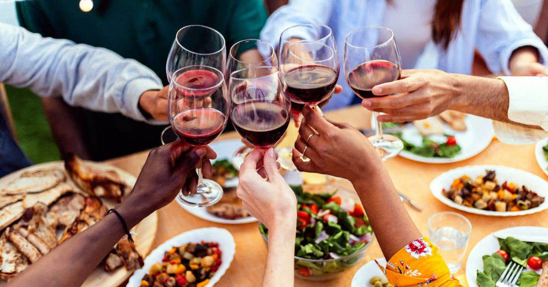 People clink glasses of red wine around a wooden dining table covered in plates of roasted vegetables and other food.