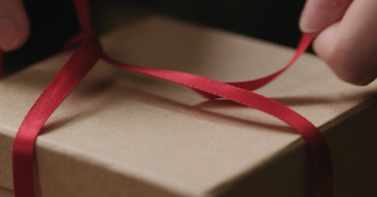 A close-up of something tying a small red, satin ribbon around a box in kraft paper. They are forming the second knot.