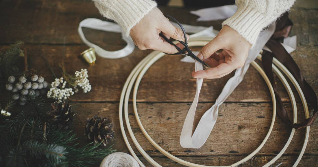 A top view of a wooden table with wreath-making supplies. Someone's hands are cutting a piece of white ribbon.