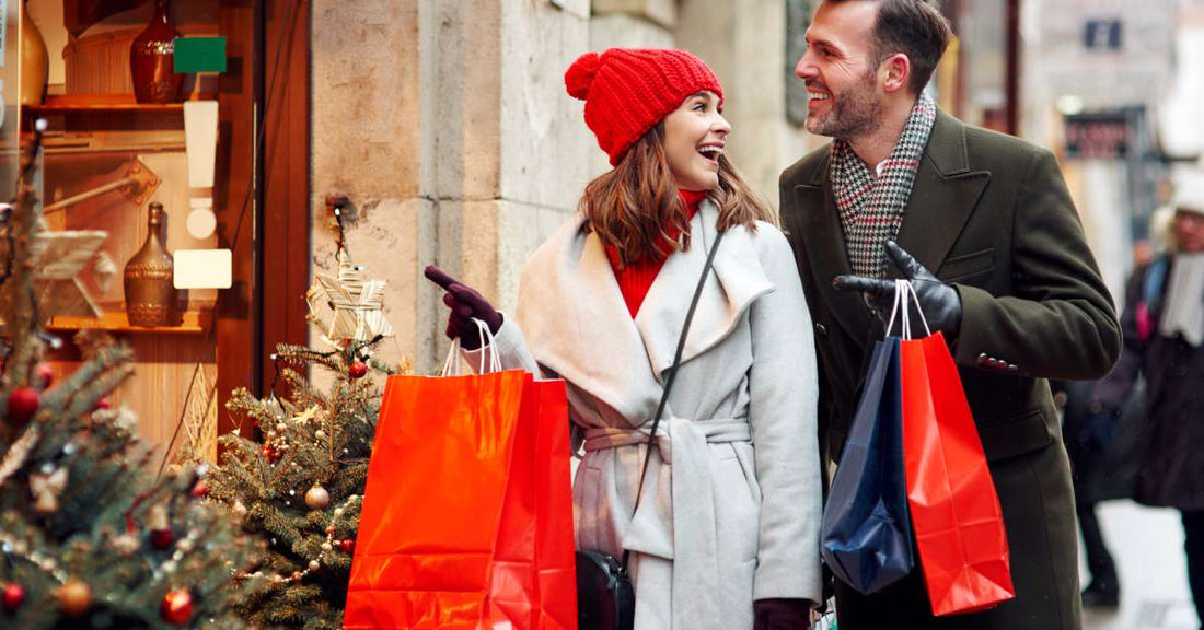 A man and woman walking down a sidewalk while holding various shopping bags. The woman points to a winter holiday store display.