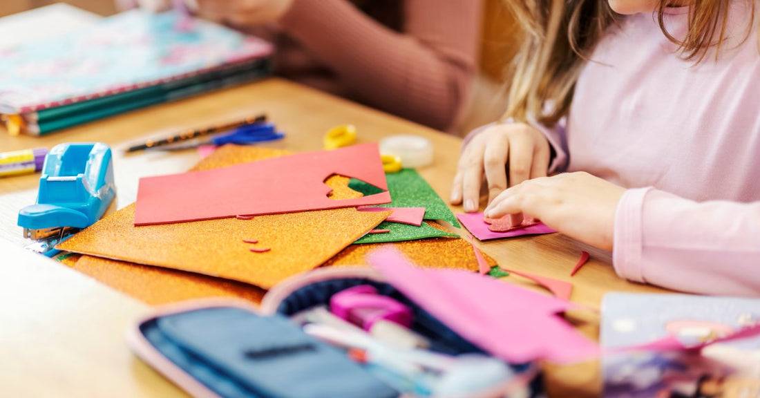 A close-up of a table with construction paper, tape, scissors, and other craft supplies. A small child is crafting something.