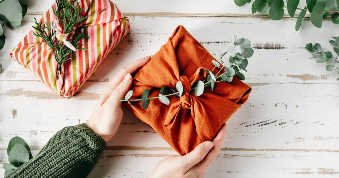 Two presents wrapped in fabric with plant stems on top. One is green, brown, and pink striped; the other is terracotta colored.