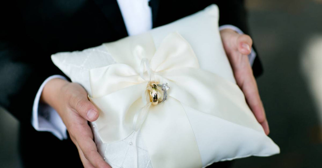 A close-up of someone holding a white ring bearer pillow. The wedding bands are wrapped in a white satin ribbon bow.