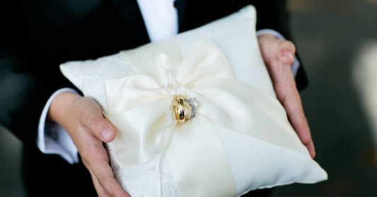 A close-up of someone holding a white ring bearer pillow. The wedding bands are wrapped in a white satin ribbon bow.