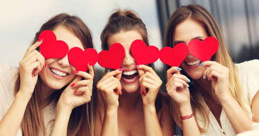 Three women sit together and have big smiles while they hold cut-out red paper hearts over their eyes.