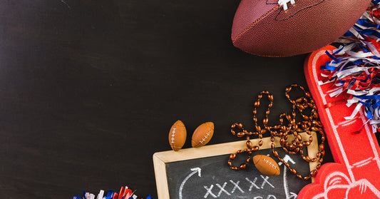 A chalkboard with a football play written on it, a football, a foam finger, pom-poms, and a necklace against a black background.