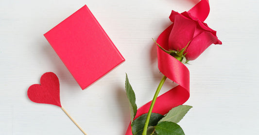 A rose with red satin ribbon twisted around it resting on a table. A heart on a stick and a small red box are next to the rose.