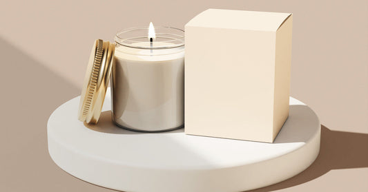 A lit white candle with a gold lid resting against it. There is a plain white box on the other side of the candle.