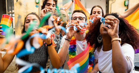 A group of people celebrate Pride by blowing on streamers. Some are also holding rainbow flags and cups.