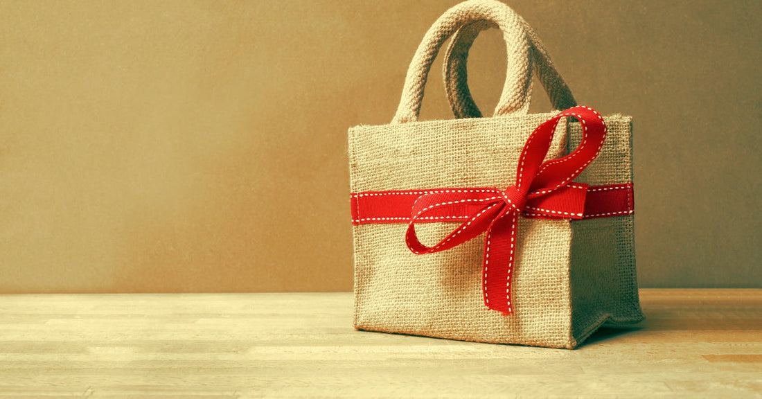 A burlap tote bag resting on a wooden table. There is a red ribbon with white stitching tied into a bow around the bag.