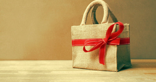 A burlap tote bag resting on a wooden table. There is a red ribbon with white stitching tied into a bow around the bag.