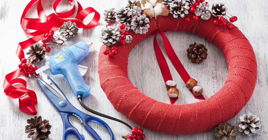 A half-completed wreath on a table. A pair of scissors, a glue gun, pinecones, and a red ribbon rest next to the wreath.