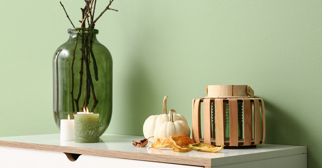 White pumpkins, leaves, candles, and branches in a vase on top of a white table. The wall is green behind the table.