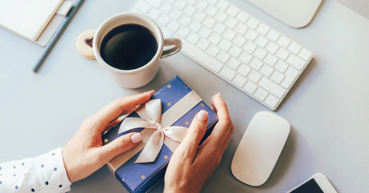 Someone sits at a desk with a gift box in their hands. A keyboard, coffee cup, mouse, and phone rest on the desk too.