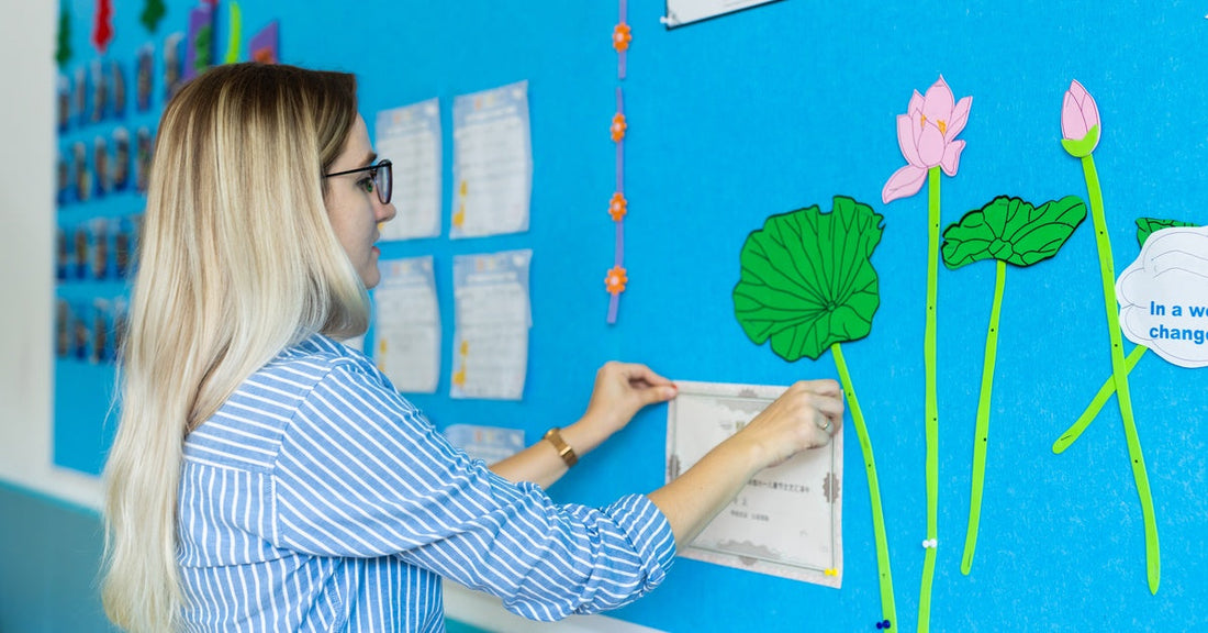 A teacher attaches a certificate to a decorated classroom bulletin board. Photos, trees, and flowers fill the board.