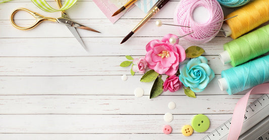 Scissors, buttons, faux flowers, paint brushes, a ruler, green gingham ribbon, and colorful threads rest on a white wooden background.
