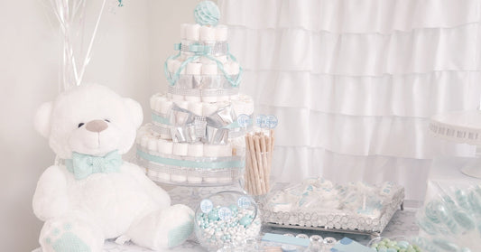 A baby shower table display with candy favors, a diaper cake, and a white teddy bear with a mint-green bow tie.