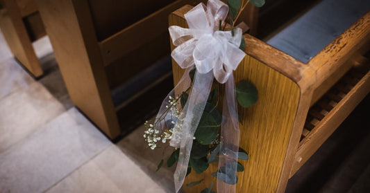 A close-up of a wooden pew with an elegant bow hanging from the side. The bow is sheer white with floral accents.
