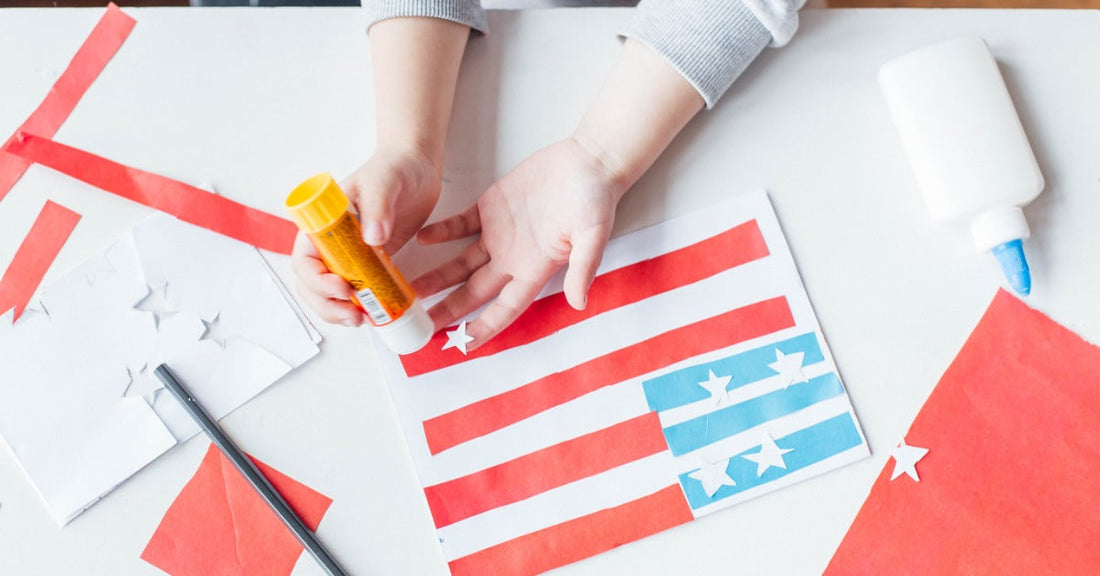 A child's small hands hold a glue stick and paste together a craft that resembles the United States of America flag.