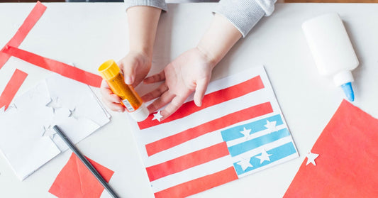 A child's small hands hold a glue stick and paste together a craft that resembles the United States of America flag.
