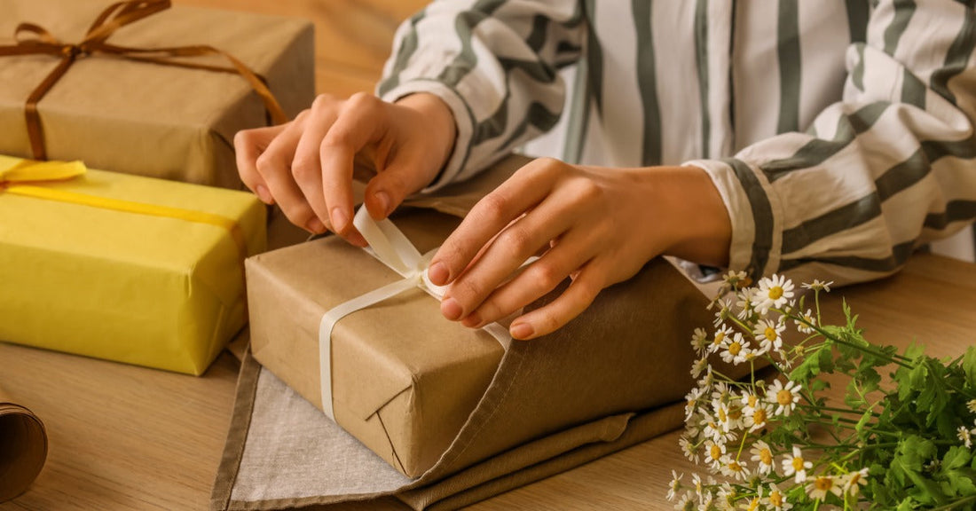 Someone tying a white ribbon into a bow on a small box wrapped in kraft paper. Flowers rest next to the package.