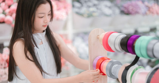 A woman stands in front of a ribbon display with many different colors. Each ribbon is on a roll and attached to a rod.